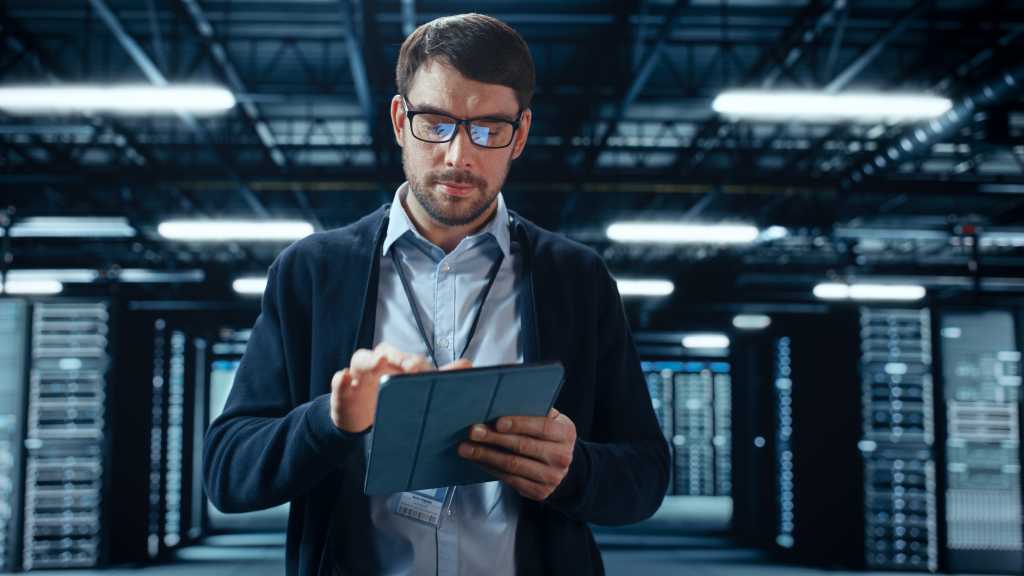 Male IT Specialist Walks Between Row of Operational Server Racks in Data Center. Engineer Uses Tablet Computer for Maintenance. Concept for Cloud Computing, Artificial Intelligence, Cybersecurity.