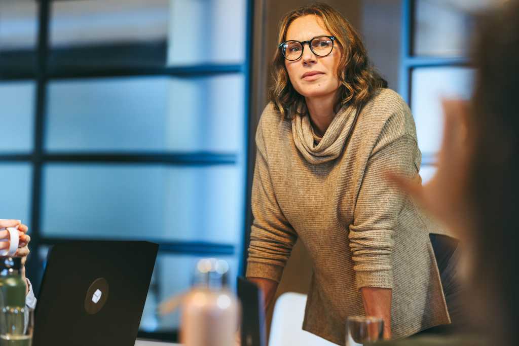 Businesswoman addresses colleagues in a boardroom. They engage in a creative discussion, promoting collaboration and teamwork. The mature leader fosters a tech-centric, startup atmosphere.