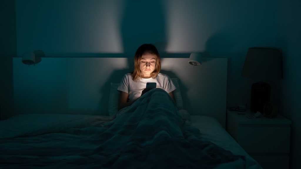 A woman sitting in bed in the dark, with her face illuminated by a smartphone in her hands.