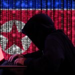 Hacker in a dark hoody sitting in front of a notebook with digital north korean flag and binary streams background cybersecurity concept