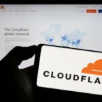 A photograph of a person holding a smartphone with the Cloudflare logo displayed. Behind the phone is a blurred monitor displaying the Cloudflare website.,