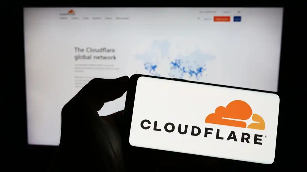 A photograph of a person holding a smartphone with the Cloudflare logo displayed. Behind the phone is a blurred monitor displaying the Cloudflare website.,