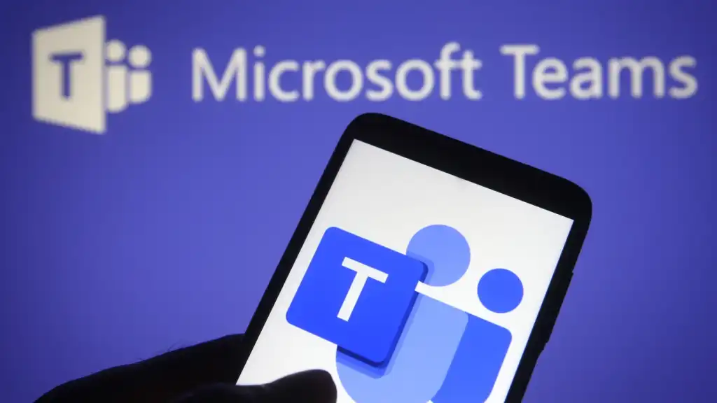 A photograph of someone holding a smartphone with the Microsoft Teams icon on it. In the background is a slightly blurred image of the Microsoft Teams logo.