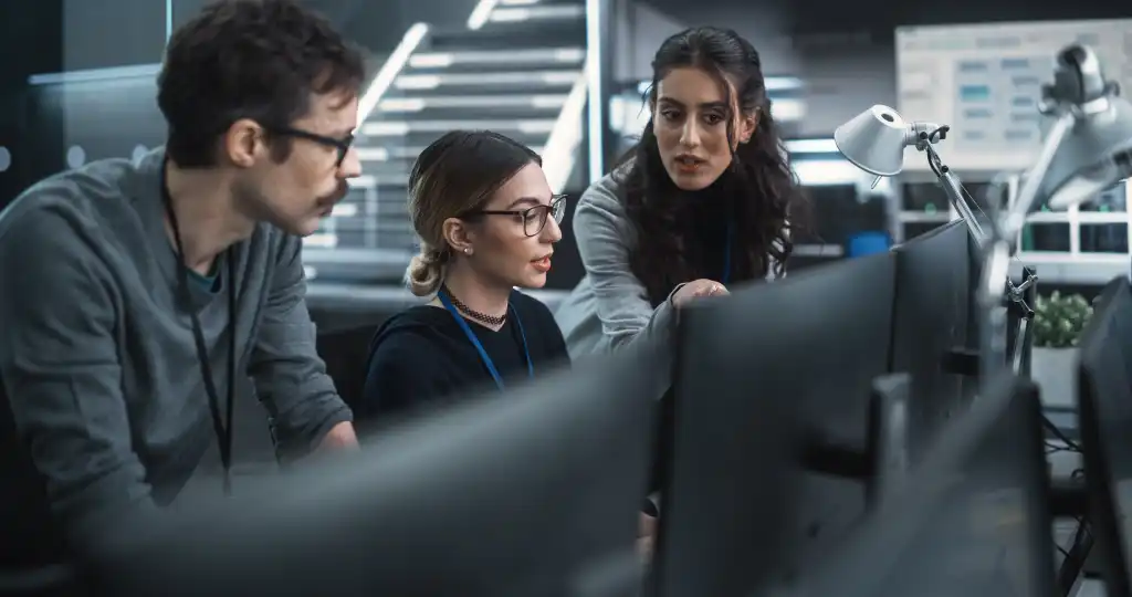 Team of Three Talented Young Software Engineers Use Computer to Discuss a Technological Project in Modern Industrial Office. Group of Male and Female Scientists Work in Research and Development Center