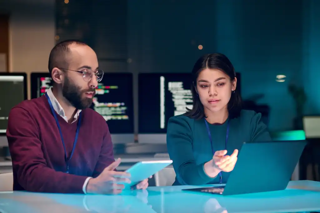 Portrait of young woman as female IT security expert using laptop with male colleague in blue neon light