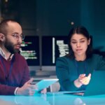 Portrait of young woman as female IT security expert using laptop with male colleague in blue neon light