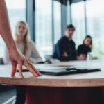 Female manager standing at table and addressing her team sitting in meeting room. Focus on hand of businesswoman in table while talking with colleagues in boardroom.