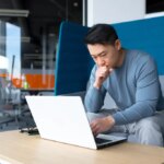 A thinking Asian man works at a computer in a modern office, a freelancer is focused on solving a task, makes an important job.