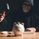 Black hat hacker in mask and hoodie trying to destroy financial piggy bank from digital bank customer dark background. Cyber security and Cybercrime concept. Business investment embezzlement theme.