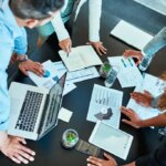 Notebook, hands and group of business people planning solution, discussion or kpi in office meeting. Laptop, sales manager and team together with profit data, budget increase or performance metrics