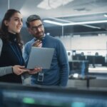 Portrait of Two Happy Female and Male Engineers Using Laptop Computer to Analyze and Discuss How to Proceed with the Artificial Intelligence Software. Casually Chatting in High Tech Research Office