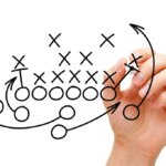 A hand draws football plays on a clear screen in black marker.