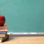shutterstock 17894746 red apple on stack of books on wood desk in front of green chalkboard