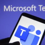 A photograph of someone holding a smartphone with the Microsoft Teams icon on it. In the background is a slightly blurred image of the Microsoft Teams logo.