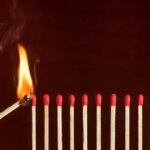 A lit match lights the first in a row of unlit matches. Lit match next to a row of unlit matches. Red phosphorus matches on dark red matte background.