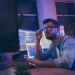 Profile photo of a developer / programmer reviewing code on monitors in his workspace.