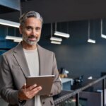 Portrait of confident mature business man executive standing in corporate office using digital tablet. Middle aged professional businessman manager wearing suit posing at work in modern space.