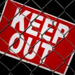 Close up view of red keep out sign with white letters behind chain link fence. Dark background.